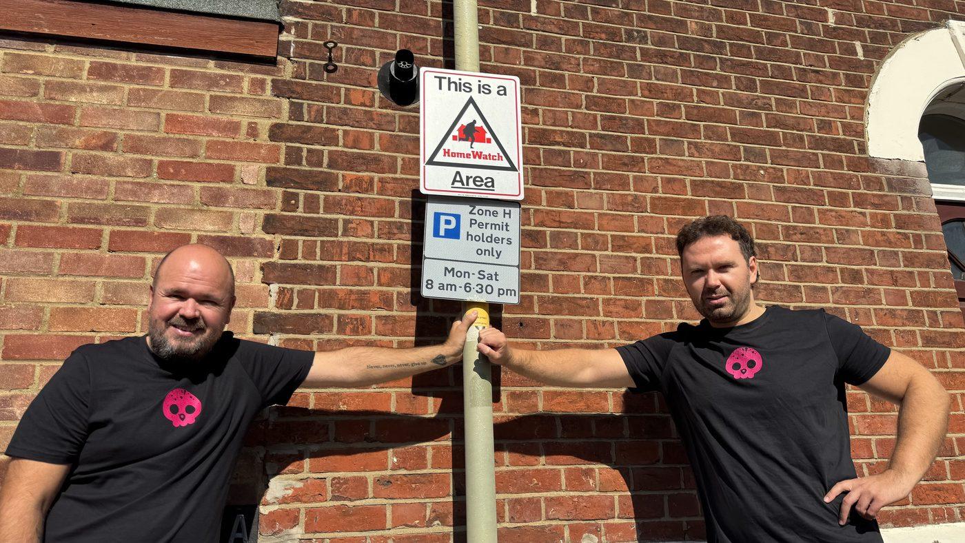 The two co-founders either side of a lamp post carrying a Home Watch area sign and a residents-only parking permit sign.